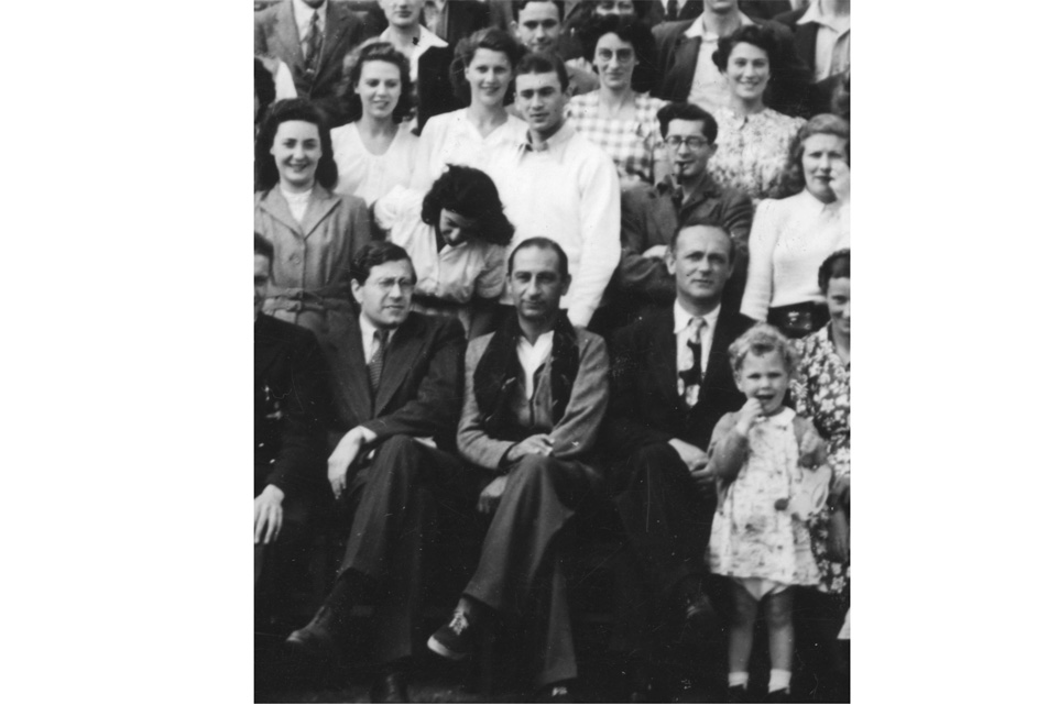 A black and white photo of a group of people wearing suits and checked dresses, with three men sitting in the front row.